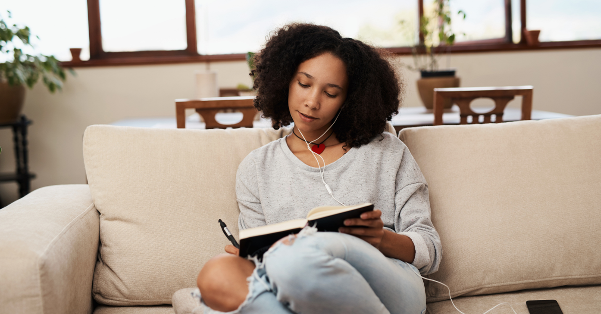 A woman sits writing in a journal with headphones in her ears.