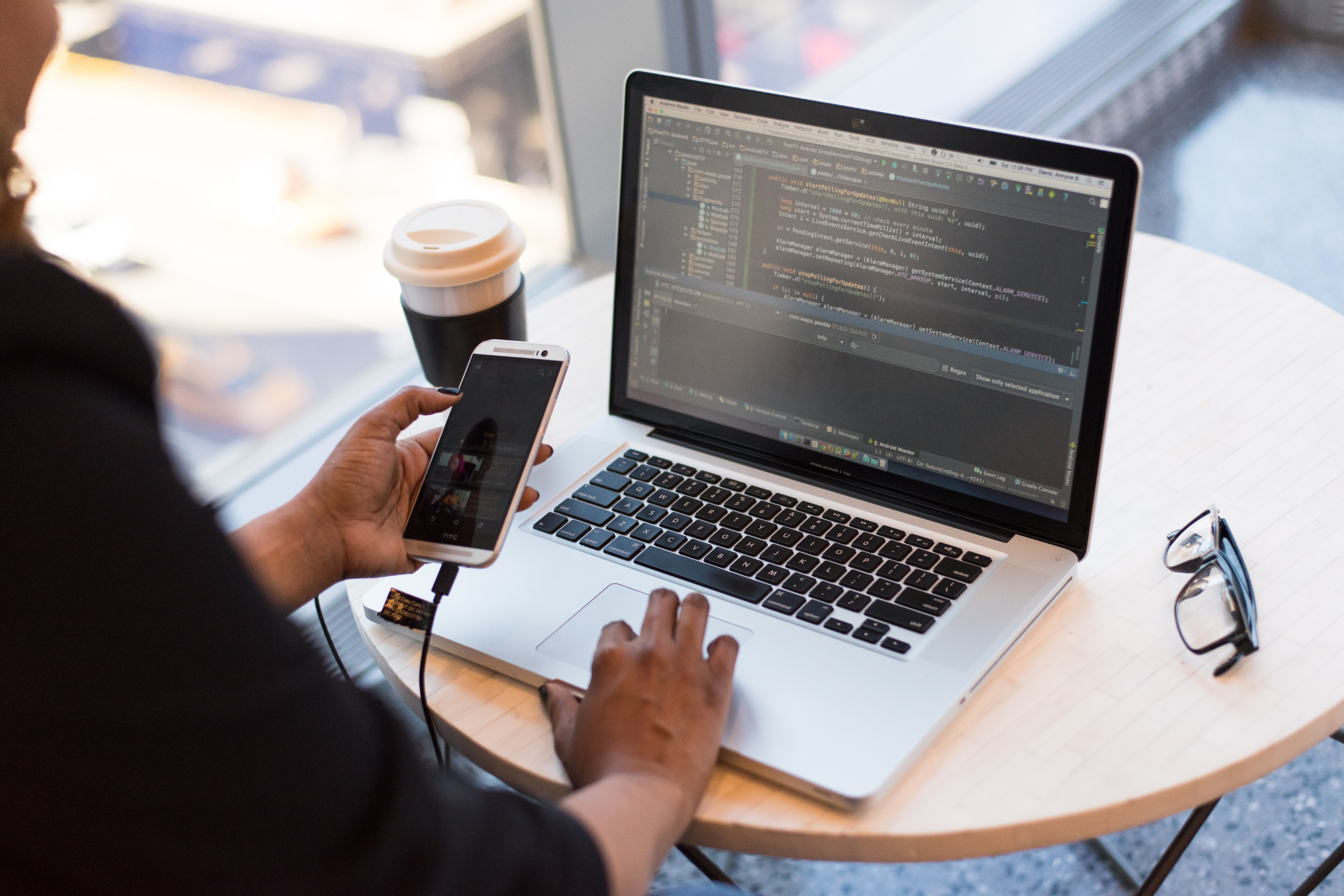 A person holds a cell phone while sitting at a table. On the table is a pair of glasses, a cup of coffee, and a laptop with some kind of technical data on it.