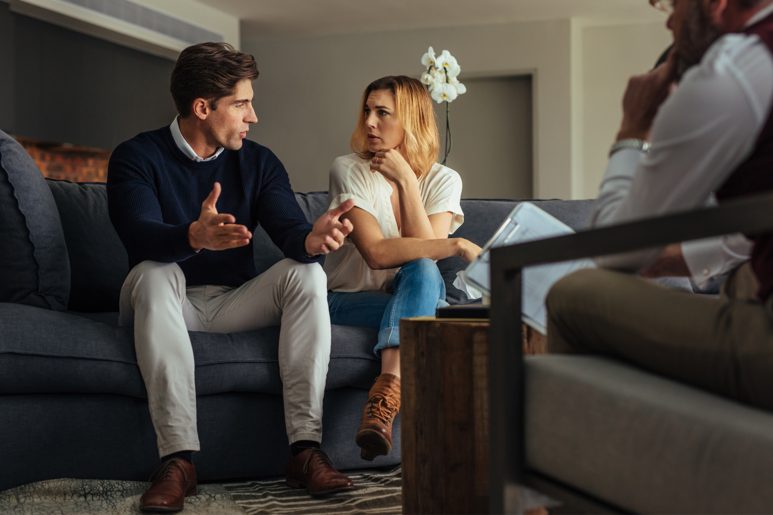 A man and woman sit next to one another in a deep conversation while a therapist can be partially seen.