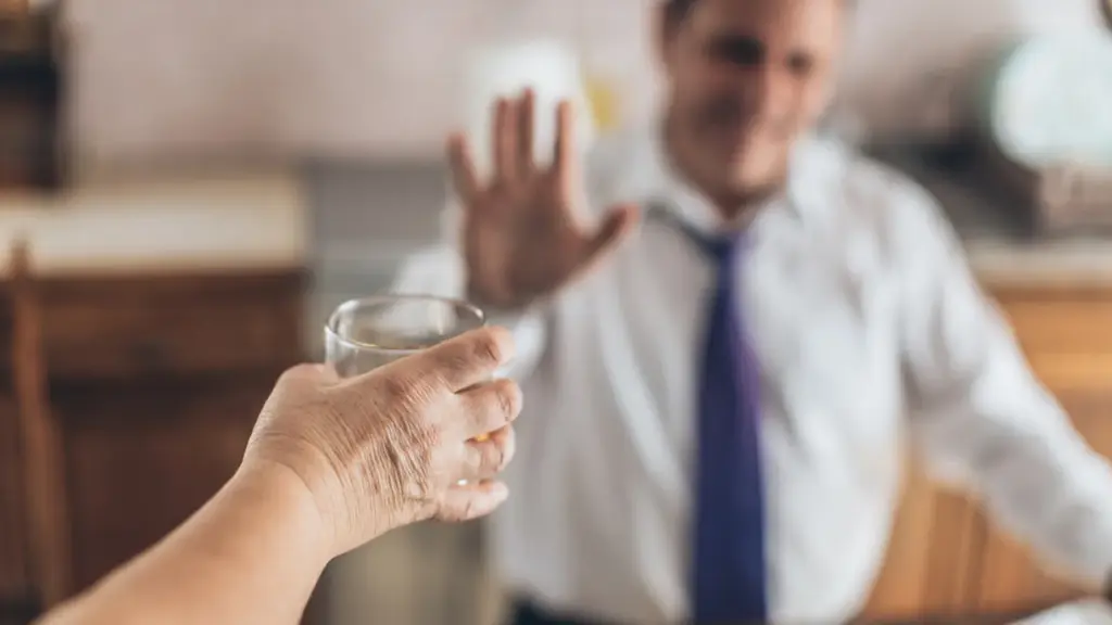 A hand is holding a glass up to the blurred image of a man who is holding up his hand in refusal.