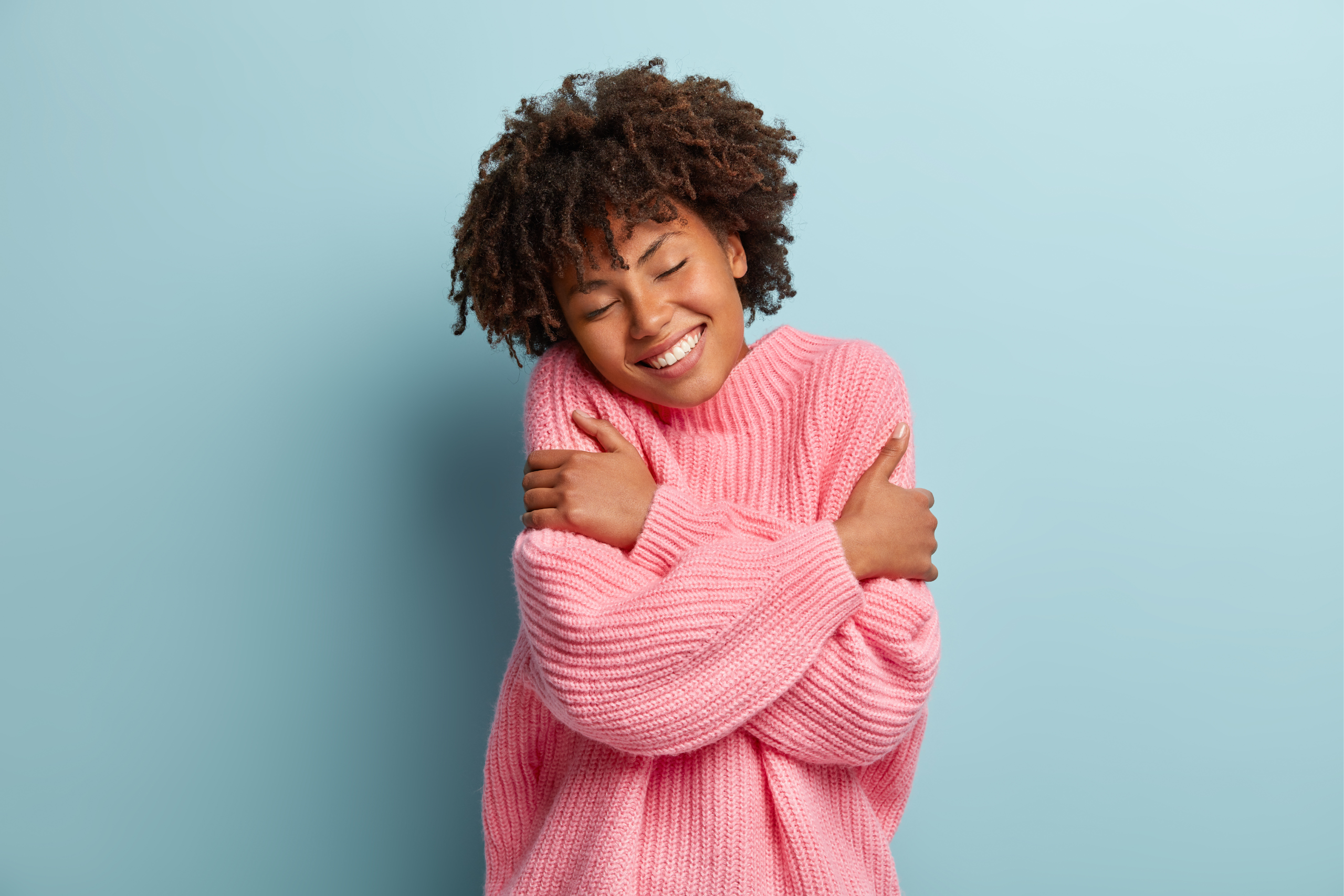 A young woman in a pink sweater smiles while giving herself a hug.
