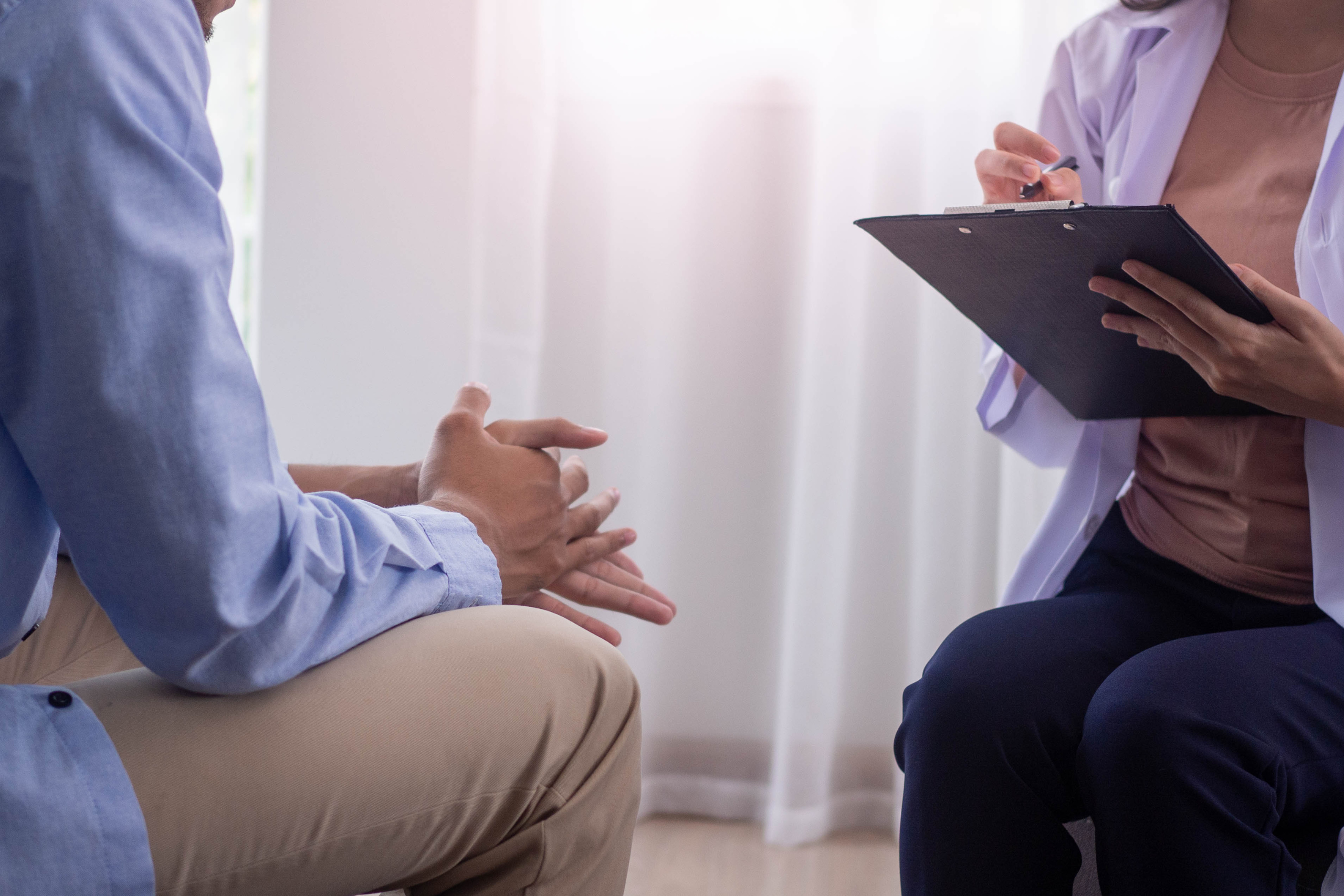 Without showing faces, one person holds a clipboard while the other clasps their hands together. They are sitting across from one another.