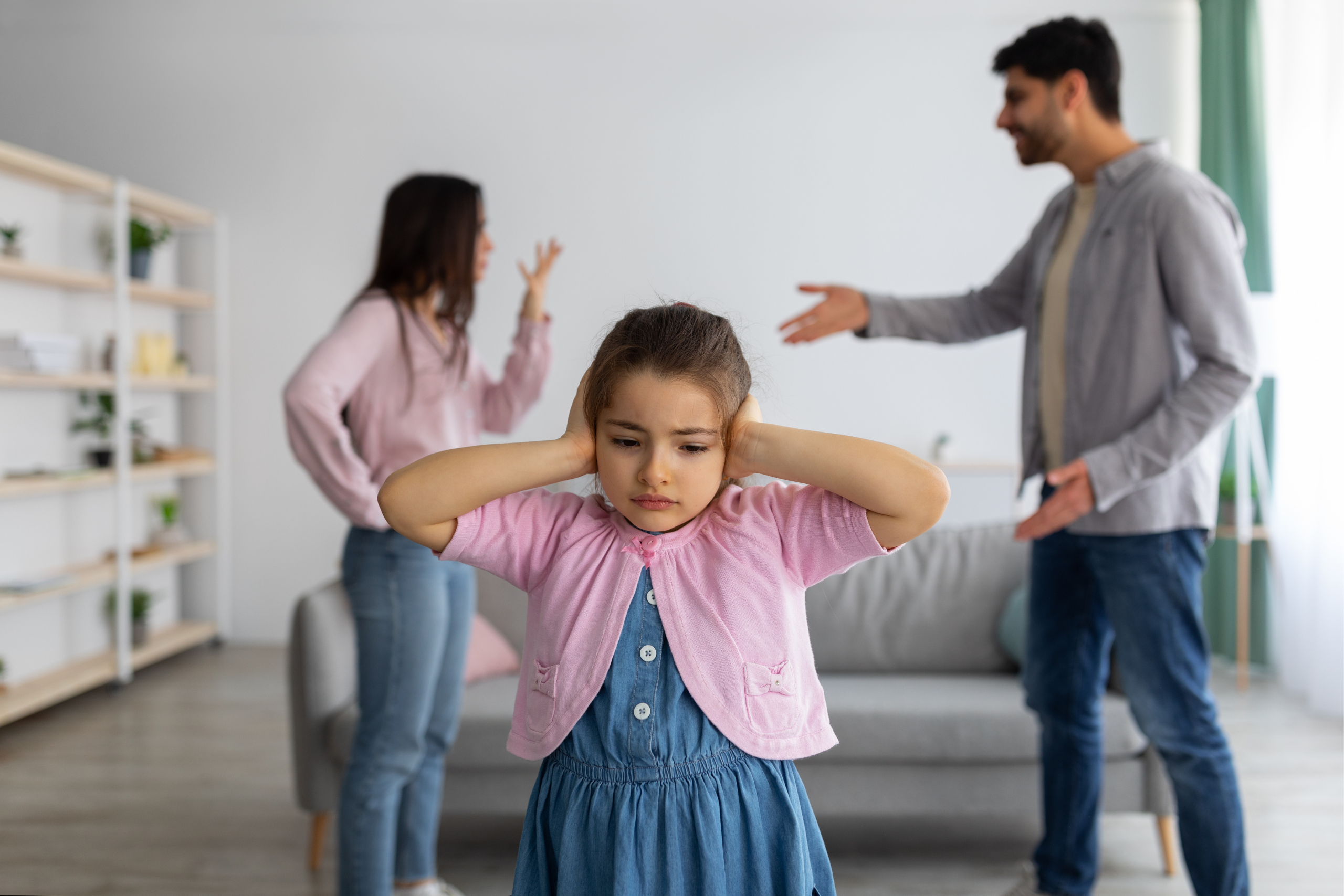 A little girl holds her ears with a sad expression while her parents argue in the background.