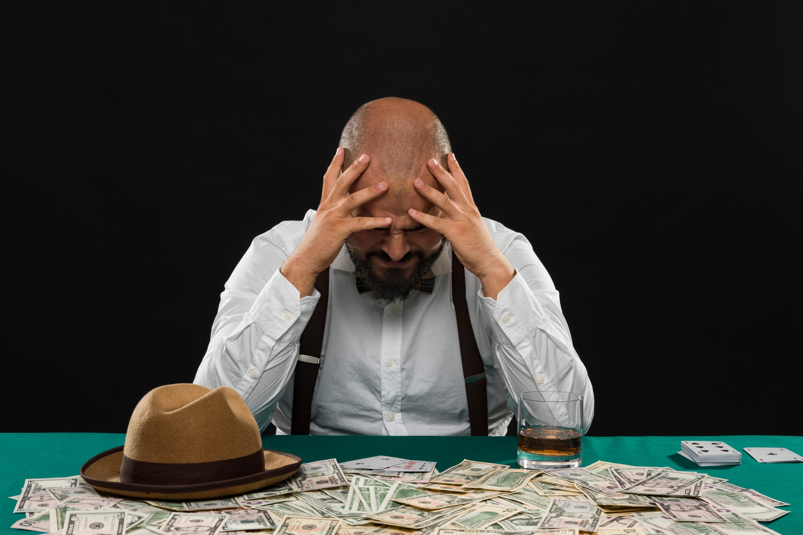 A man sits with his hat off at a poker table with a pile of cash. He is hold his head as if he was stressed.