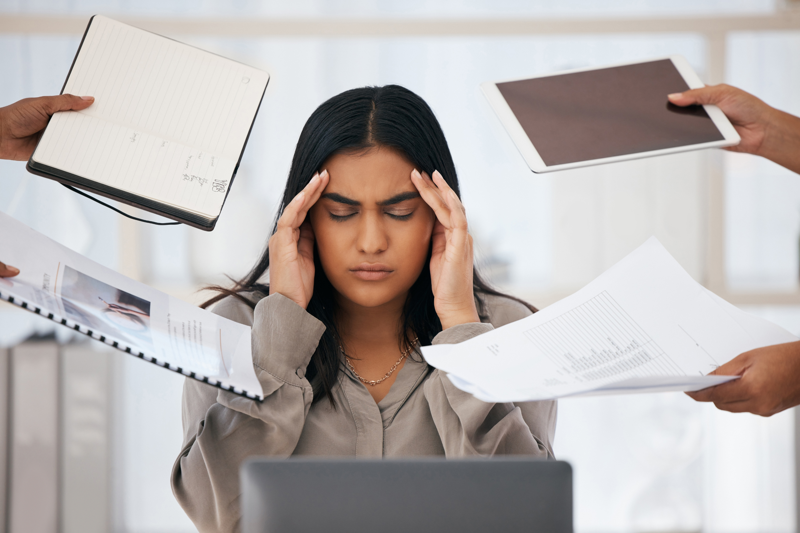 A woman holds her temples in stress while several hands hold different documents up to her face.