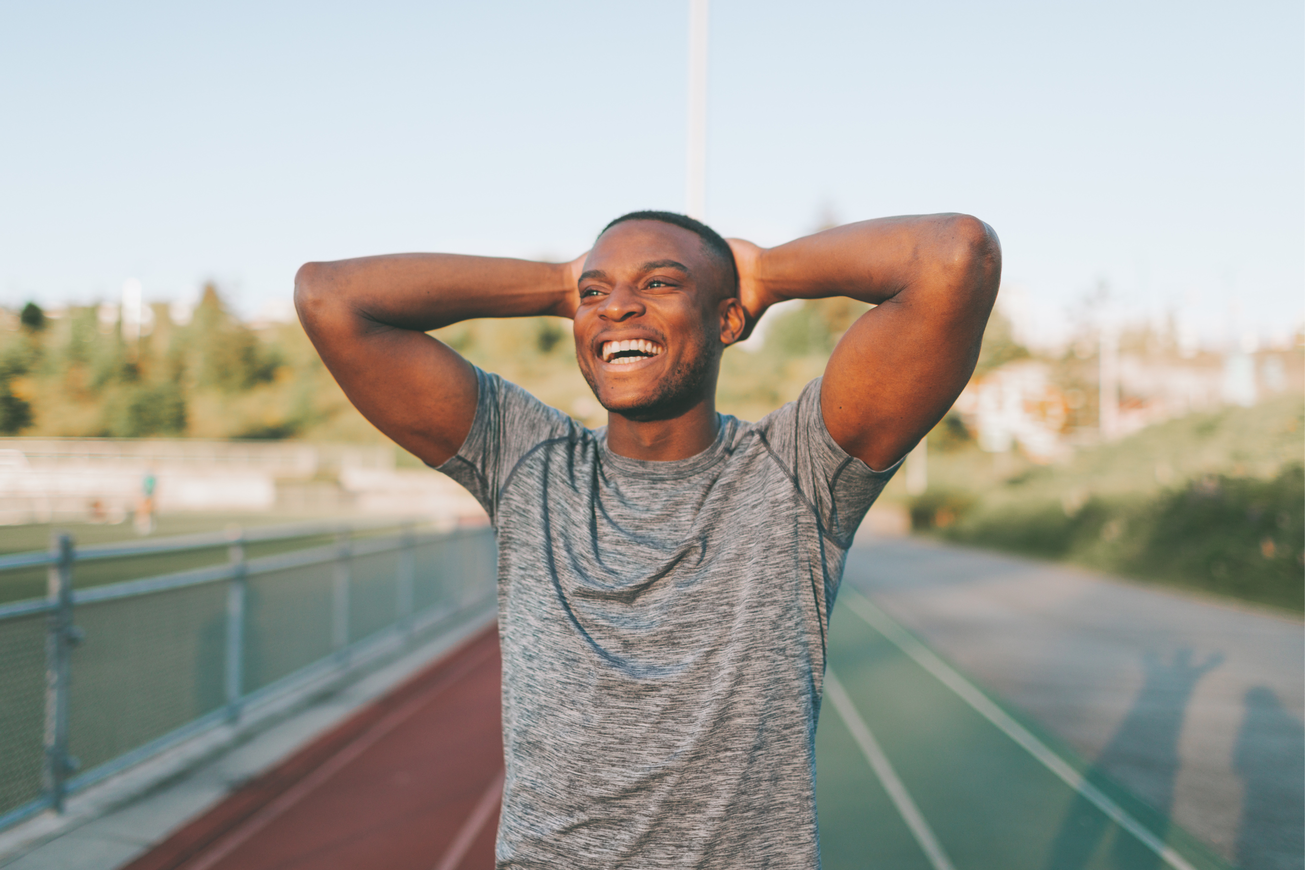 A man at a trackfield stands with his hands on the back of his head and a big smile on his face.