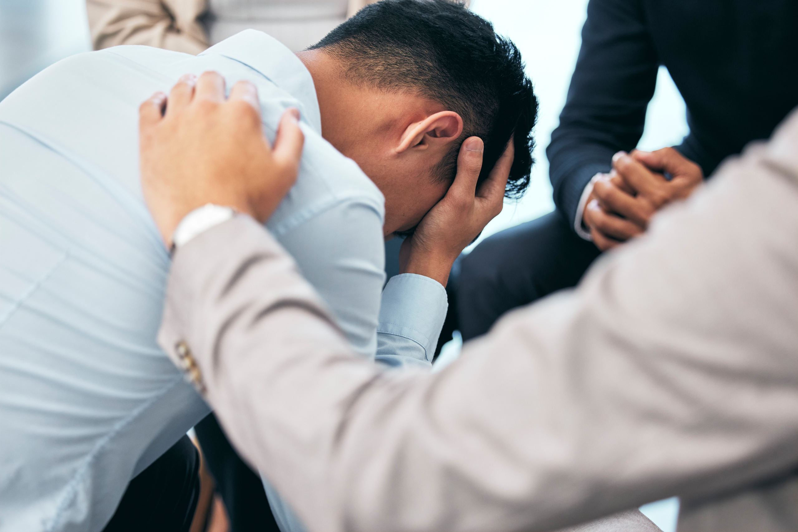 Distressed man with his head in his hands, surrounded by supportive people, symbolizing emotional pain and the importance of support during a mental health relapse.