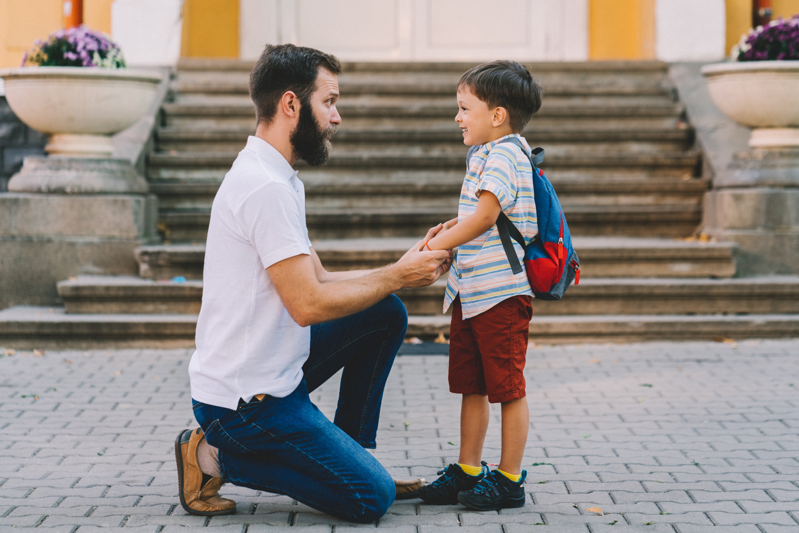 A father kneels in front of his son, holding his hands while the child smiles back at him.