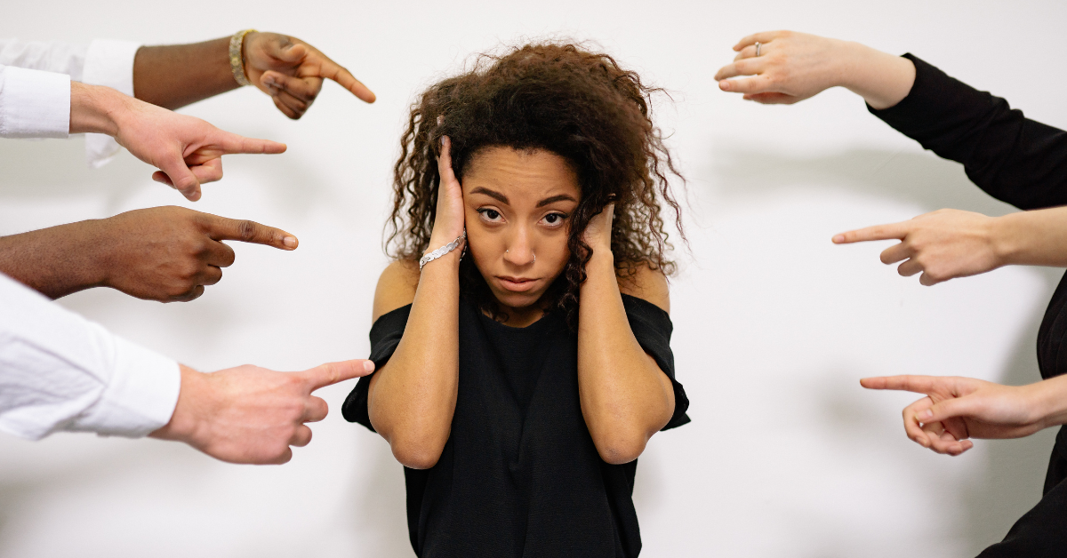 A woman stands with her hands on her ears while several hands point at her.