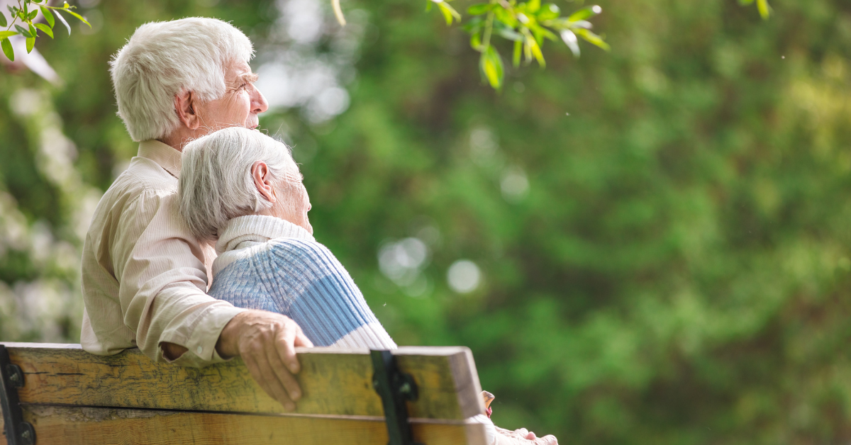 An older couple sit next to one another on a bench, facing away. The woman leans into the man.