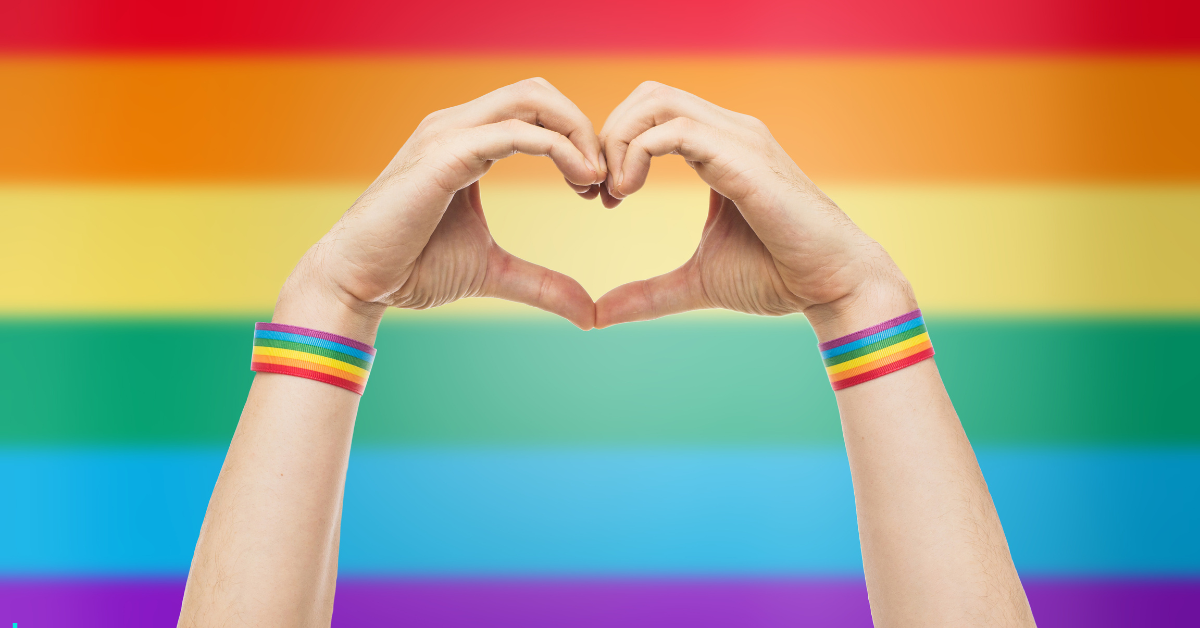 A person with hands forming a heart shape in front of a rainbow flag.