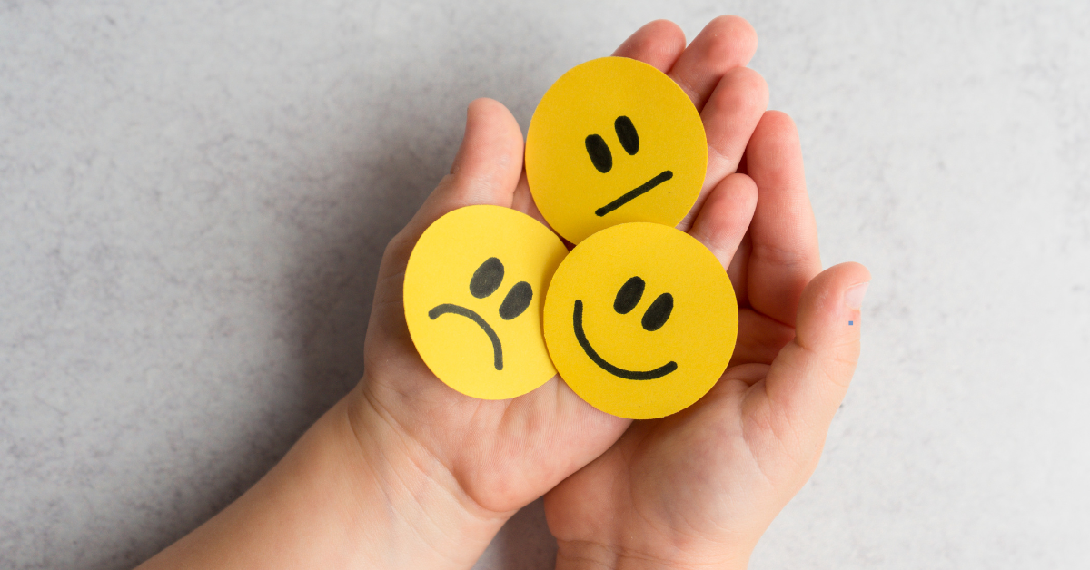A person holding three emoji faces in their hands. The emoji faces are yellow and show different emotions: happy, sad, and neutral.