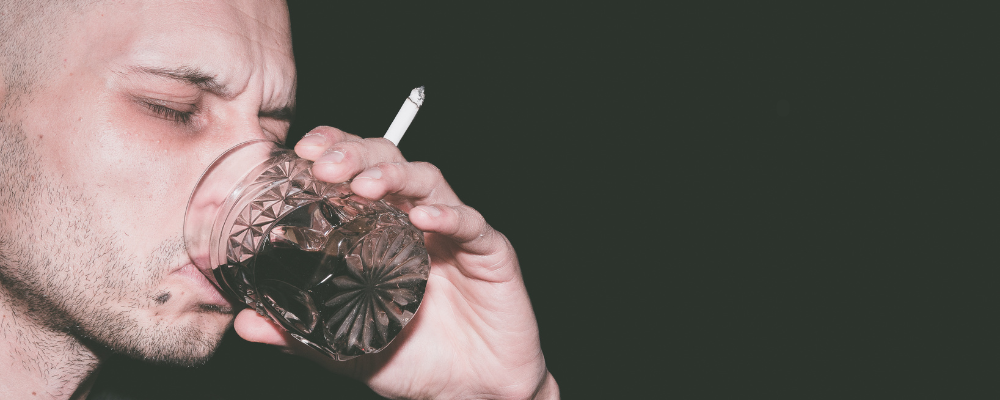 Closeup of a man drinking from a glass with a cigarette in his hand.