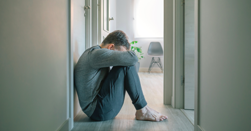 sad man in a gray sweater sitting on the floor with his head on his knees