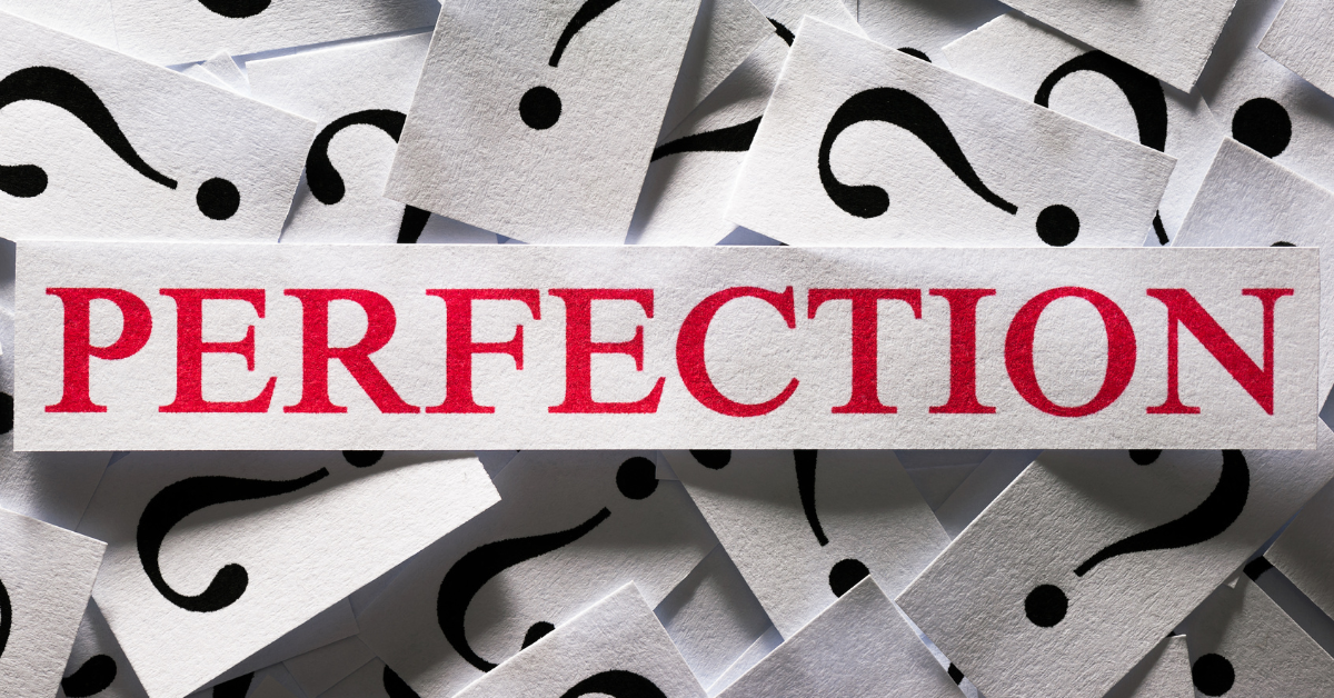 The word "PERFECTION" written in red letters on a strip of paper surrounded by question marks on white pieces of paper.