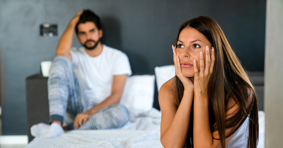 Woman sitting at the edge of the bed with her head in her hands. Her partner sits behind her up against the bed frame.