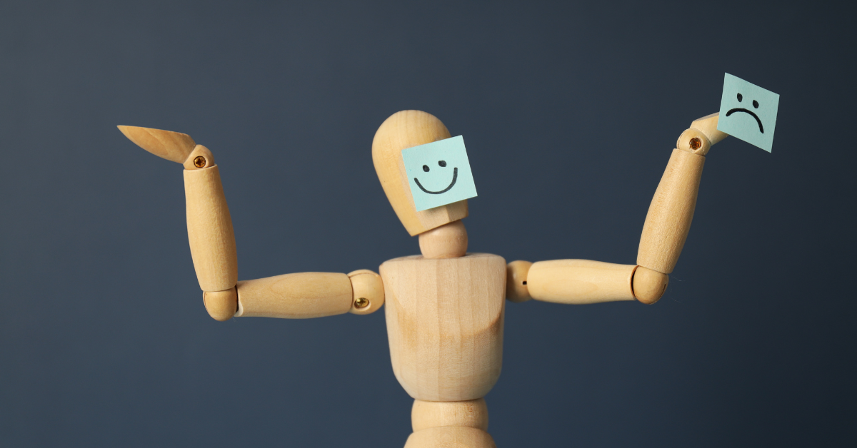 Wooden mannequin holding up two sticky notes, one with a smiling face and the other with a sad face, representing contrasting emotions and mood swings.