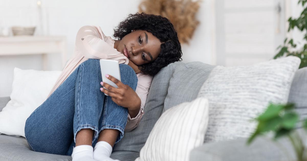 A woman sits curled up on a couch, looking at her phone with a distant and contemplative expression.