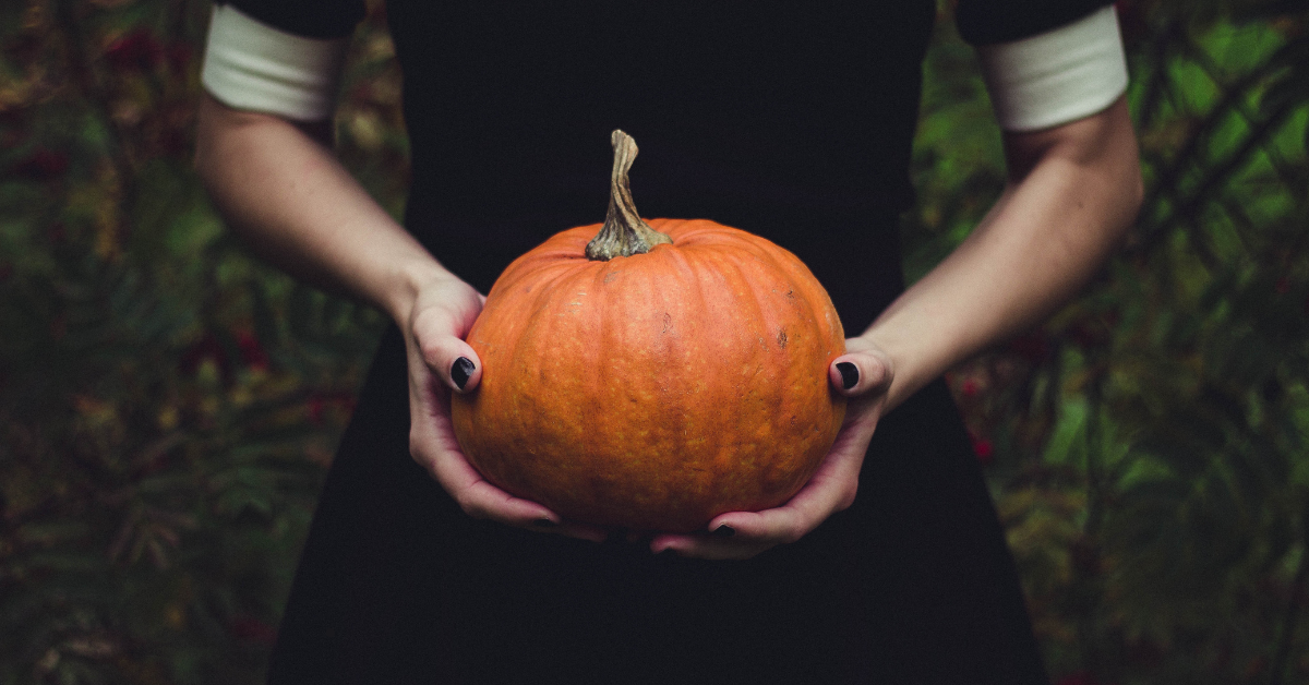A woman in a black dress with black nails holding a pumpkin.
