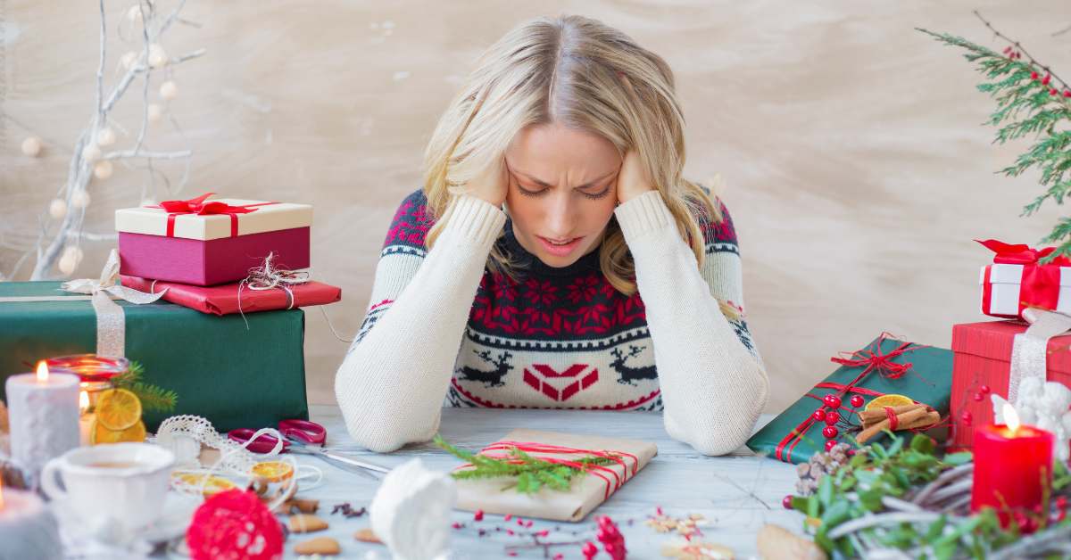 A woman in a festive sweater, looking stressed and holding her head in her hands, sits at a table surrounded by holiday decorations and wrapped gifts.