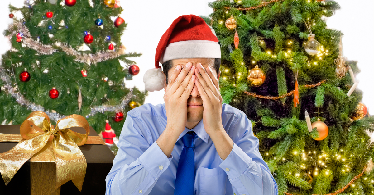 A man in a Christmas hat holds hands to his face in front of two Christmas trees.