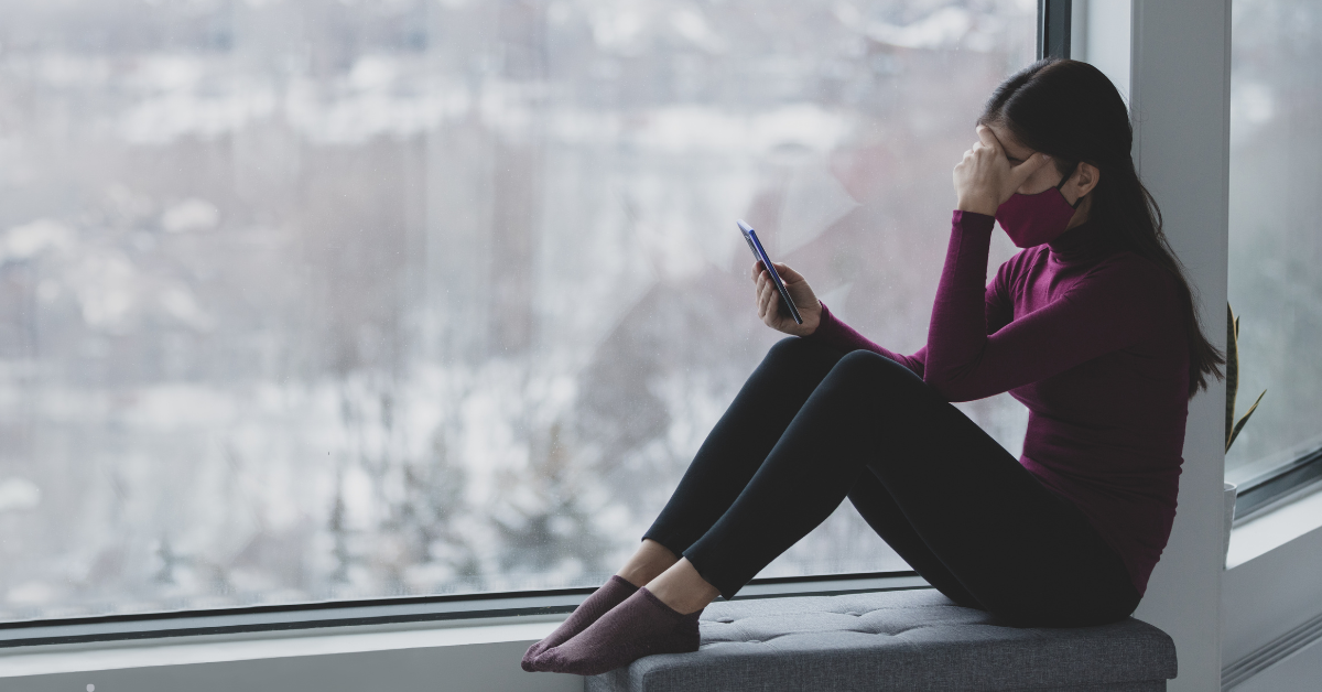 Masked woman looks at her phone sitting next to a window.