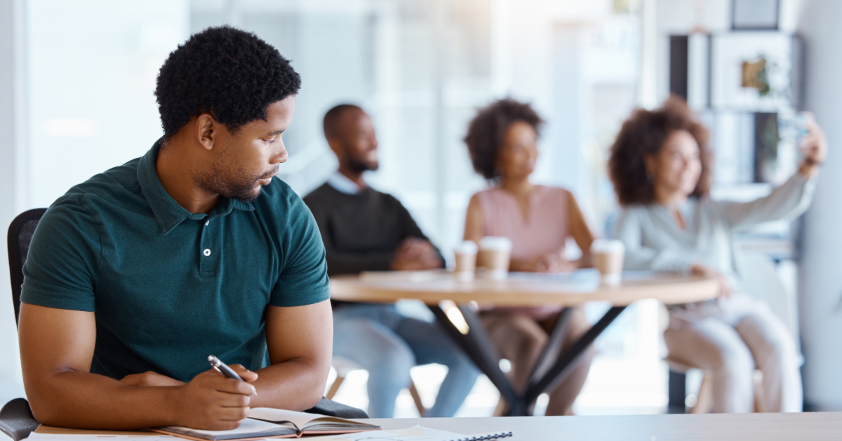 A man sits looking at a group behind him that are taking a selfie.