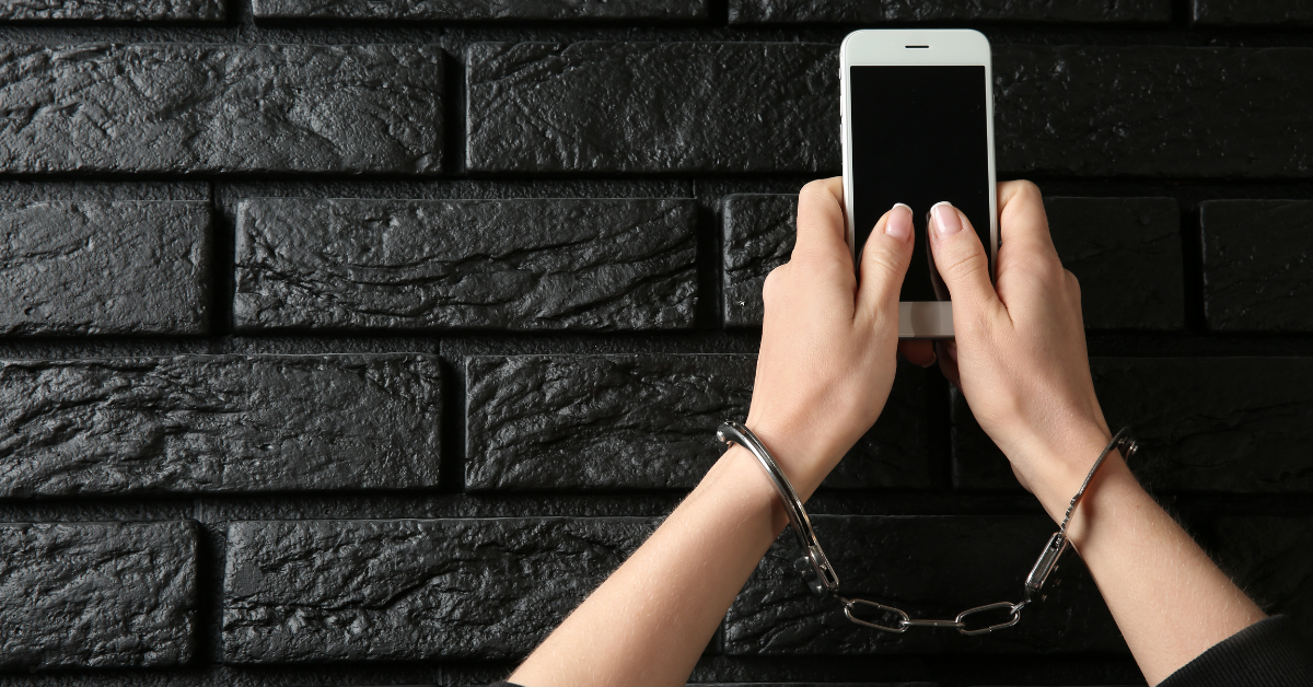 A pair of handcuffed hands holds up a smart phone against a black brick wall.