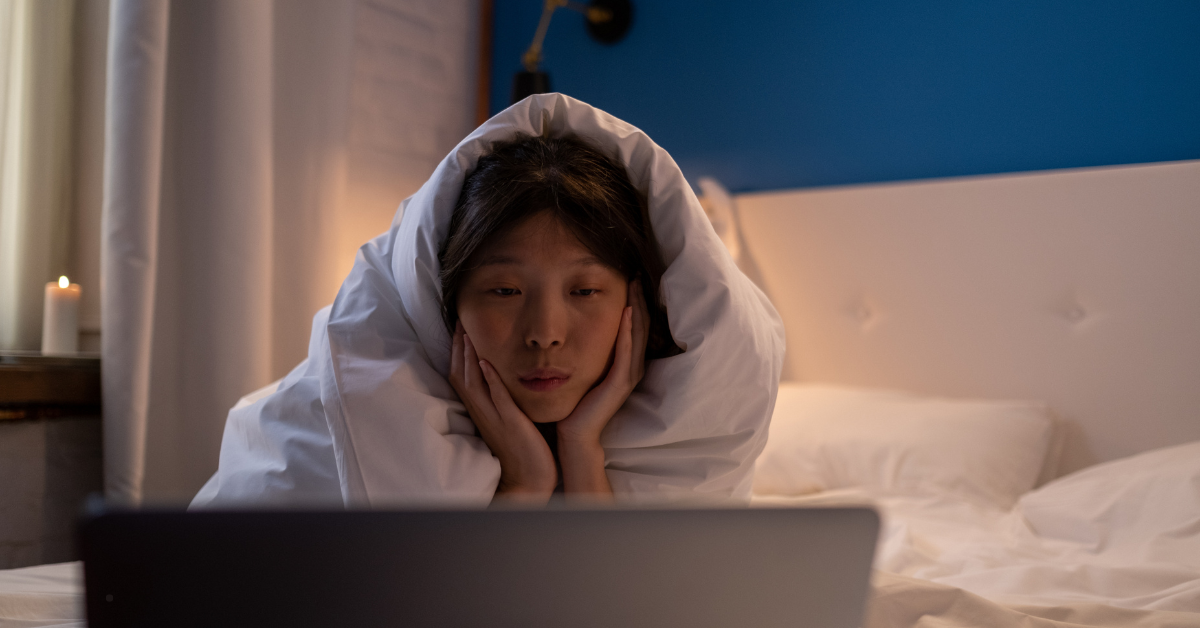 A person wrapped in a blanket sits on a bed, gazing intently at a laptop screen in a cozy, dimly lit room.