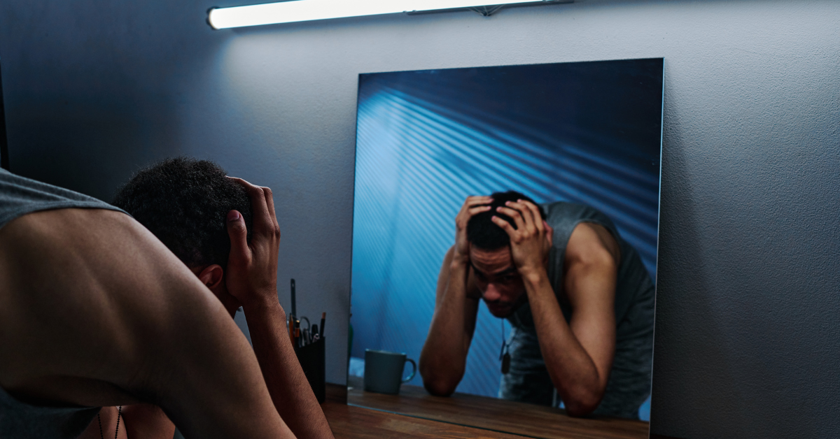 Man leaning over a desk and holding his head in frustration, reflected in a mirror under dim lighting.