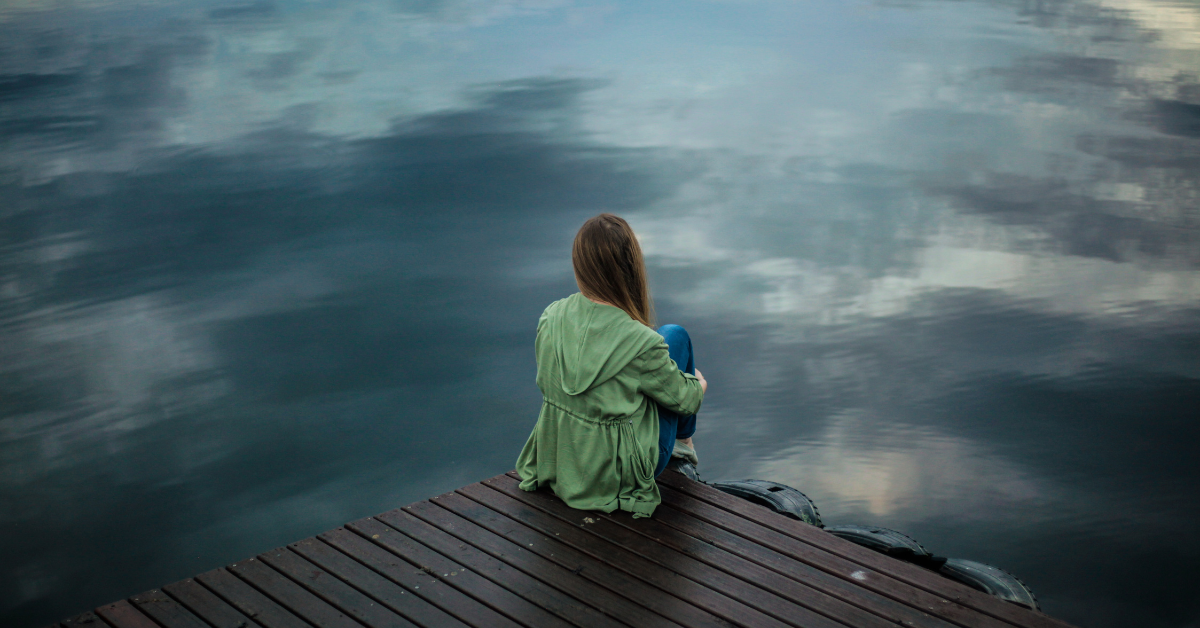Person sitting alone on a wooden dock, gazing at a calm and reflective body of water under a cloudy sky.