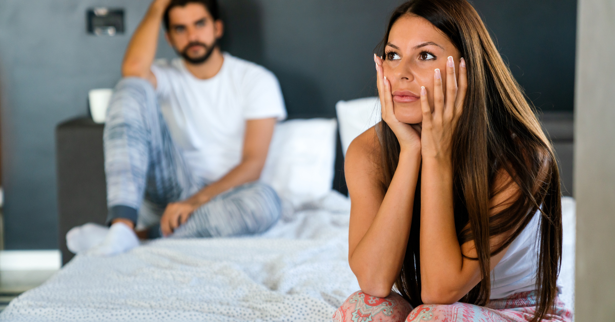 Couple sitting in a bedroom looking upset and distant, with the woman appearing contemplative and the man in the background holding his head.