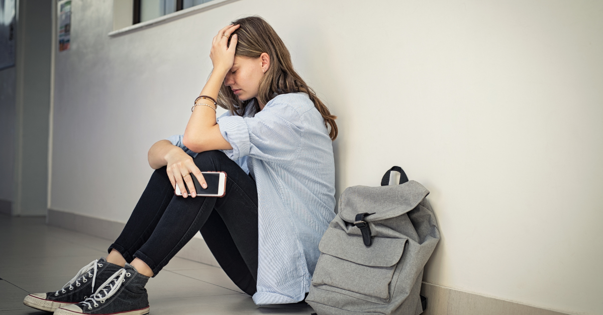A young woman sits on the floor in a school or college hallway, leaning against a wall with her head resting on her hand. She looks distressed or overwhelmed, holding a smartphone in her other hand. A gray backpack sits beside her.
