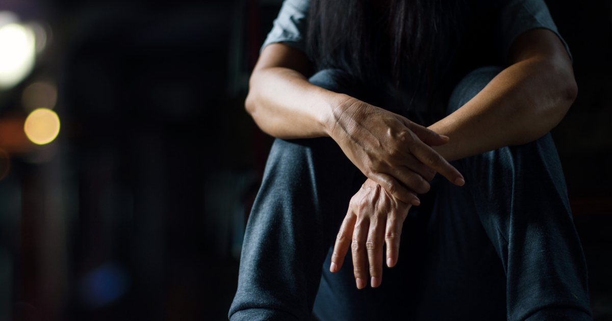 Close-up of a person sitting with arms crossed over knees in a dimly lit space, conveying emotional distress and symptoms associated with complex PTSD.