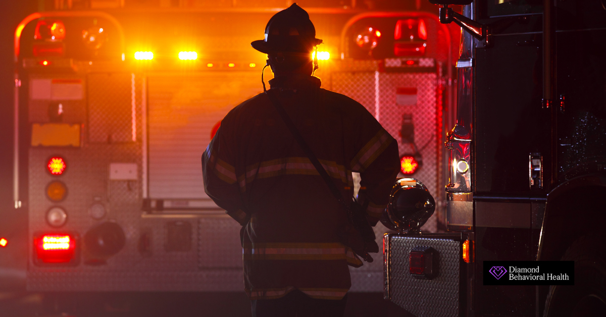 Firefighter standing between two fire trucks with emergency lights flashing, symbolizing the intensity of the profession and the mental health challenges faced by firefighters.